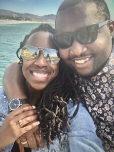 AMI employee Kevwe Odima posing with his wife for a selfie in front of in ocean on a California beach.